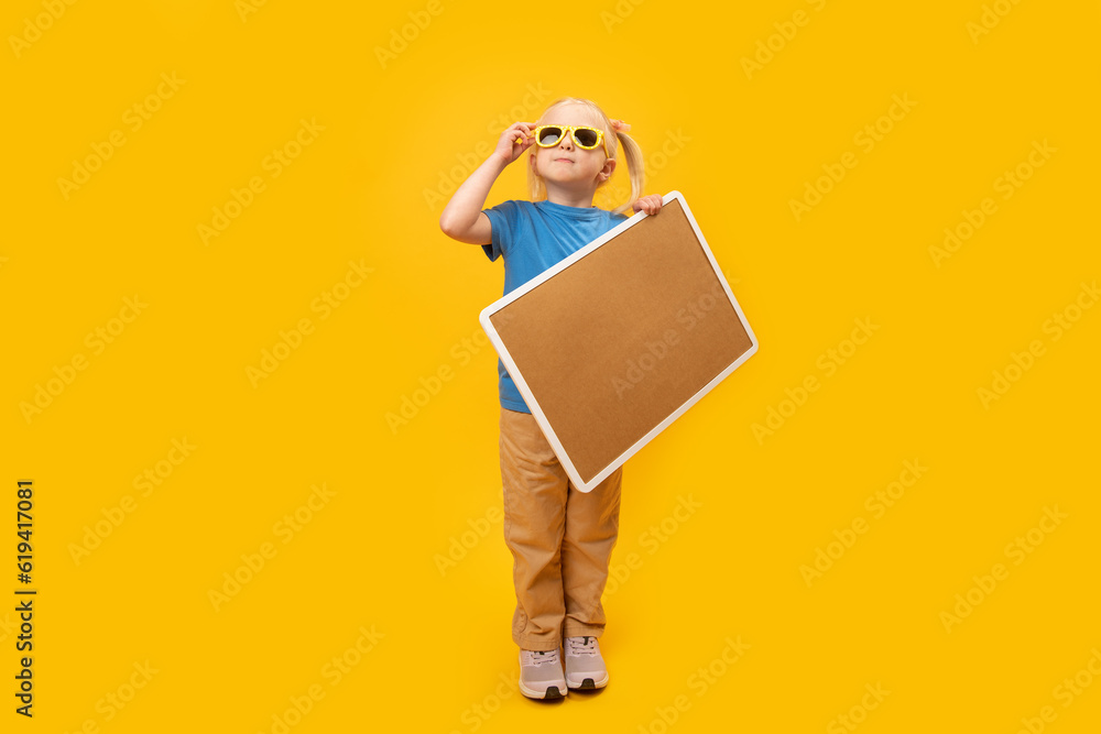 Little blonde girl with two ponytails wears sunglasses and holds cork board. Portrait of child with blank board on yellow background. Little blonde girl with two ponytails wears sunglasses and holds cork board. Portrait of child with blank board on yellow background.