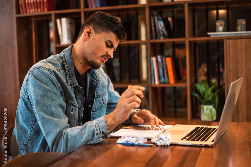 A stressed male student crumples up the paper while studying or working inside university library or home workplace. A stressed male student crumples up the paper while studying or working inside university library or home workplace.