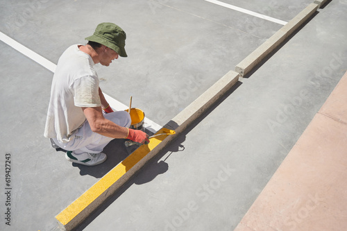 Professional painter at work. Young man uses a paint roller to apply special acrylic paint for road marking on asphalt of a parking lot. 
