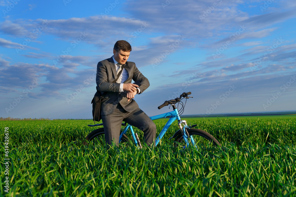 a businessman rides a bicycle with a briefcase and checks his wrist watch on a green grassy field, dressed in a business suit, beautiful nature in spring, freelance business concept a businessman rides a bicycle with a briefcase and checks his wrist watch on a green grassy field, dressed in a business suit, beautiful nature in spring, freelance business concept
