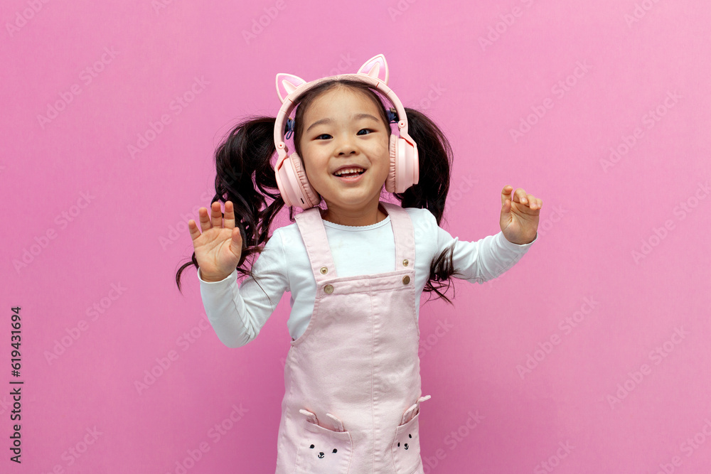 little asian girl in pink children's headphones listens to music and dances on pink isolated background, korean child little asian girl in pink children's headphones listens to music and dances on pink isolated background, korean child