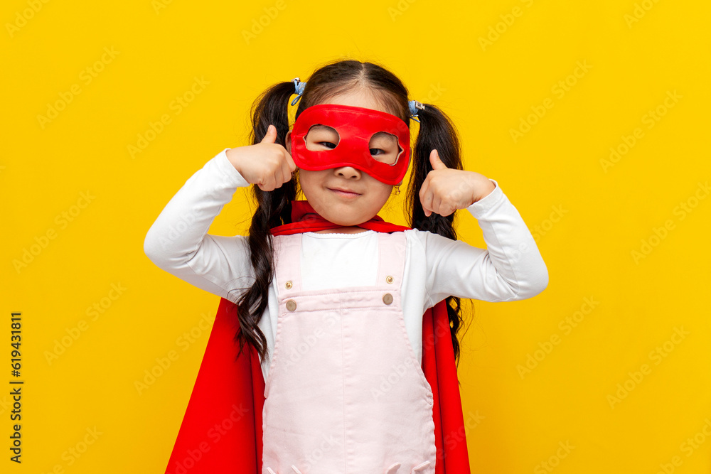 little asian girl in superman costume and mask shows likes with her hands, korean child in cape smiles little asian girl in superman costume and mask shows likes with her hands, korean child in cape smiles