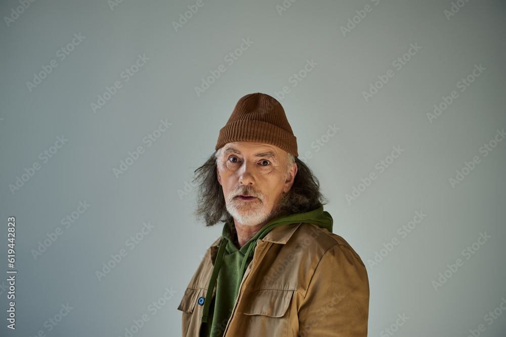 thoughtful and discouraged hipster style senior man in beanie hat and brown jacket looking at camera while standing on grey background, aging population lifestyle concept thoughtful and discouraged hipster style senior man in beanie hat and brown jacket looking at camera while standing on grey background, aging population lifestyle concept