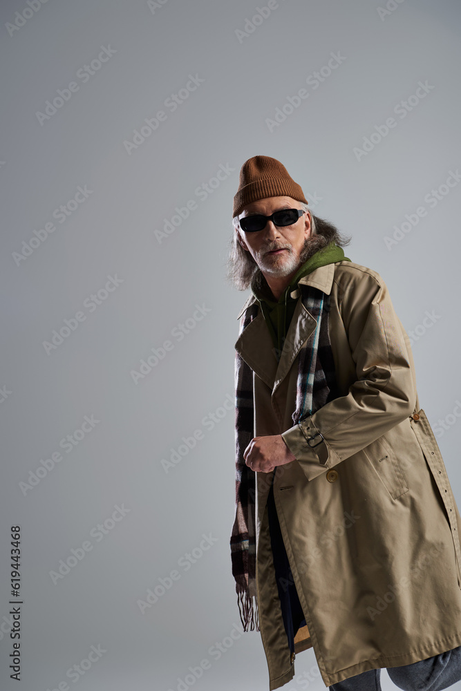 grey haired senior man in dark sunglasses, beanie hat, beige trench coat and plaid scarf looking away on grey background, expressive personality, hipster fashion, positive aging concept grey haired senior man in dark sunglasses, beanie hat, beige trench coat and plaid scarf looking away on grey background, expressive personality, hipster fashion, positive aging concept