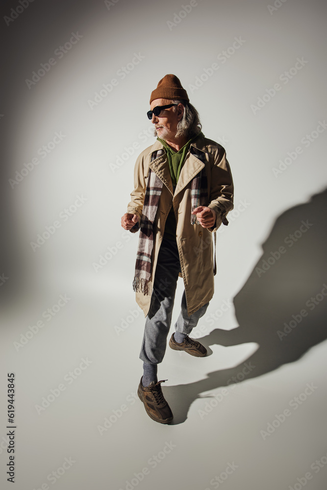 expressive personality, senior hipster man in dark sunglasses, beanie hat and beige trench coat standing in stylish pose on grey background with shadow, positive and trendy aging concept expressive personality, senior hipster man in dark sunglasses, beanie hat and beige trench coat standing in stylish pose on grey background with shadow, positive and trendy aging concept