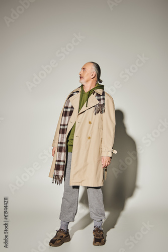 full length of senior male model in beige trench coat, plaid scarf and green hoodie standing on grey background with shadow and looking away, stylish casual attire, fashionable aging concept