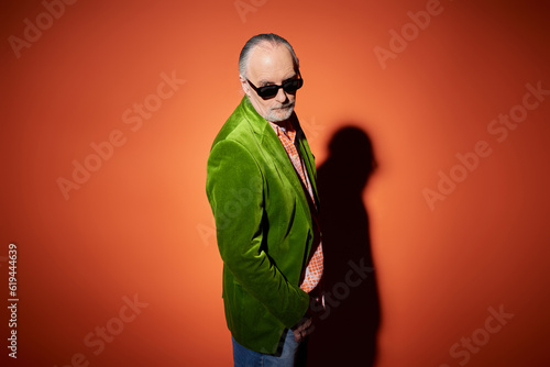 senior male model in stylish casual clothes looking at camera on red and orange background with shadow, green velour blazer, dark sunglasses, expressive individuality, fashionable aging concept
