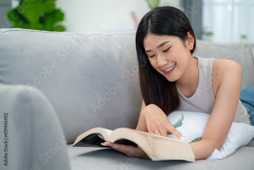 Asian woman lying on the sofa in the living room reading a book happily in the morning.