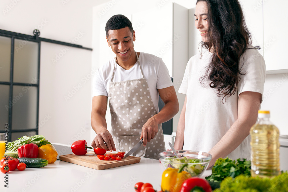 african american man in apron cuts tomatoes and looks at his girlfriend and smiles, multiracial couple prepares salad