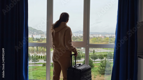 Photos Woman looks at cityscape outside panoramic window and prepares for trip holding suitcase