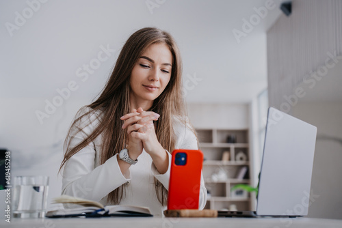 Sad blonde caucasian woman in white suit sitting at desk with laptop watching news using phone. Tired businesswoman received bas news during video call with business partner. Financial people. photo