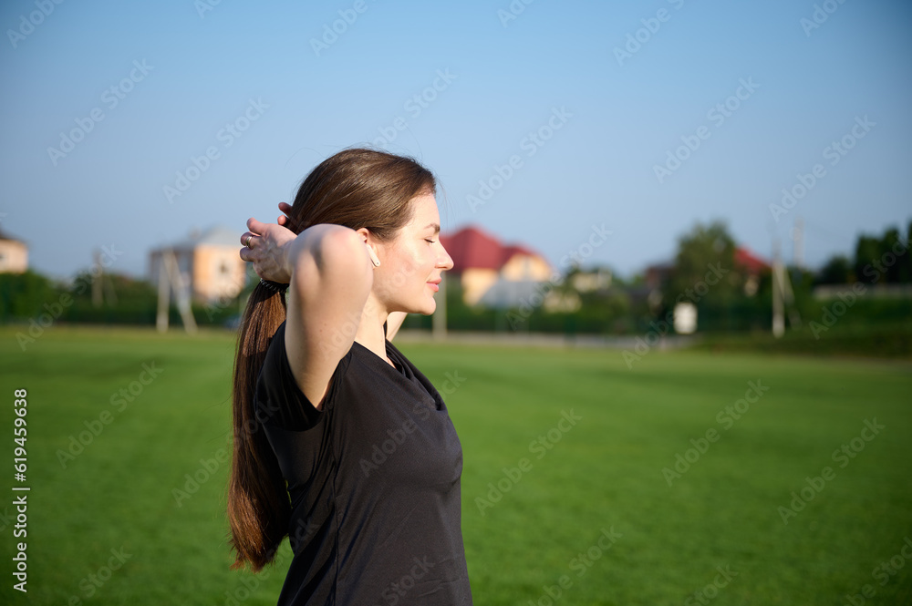 Obraz premium Portrait of young attractive fit girl in black sports tight clothes standing on green field before workout at sunrise. Caucasian female jogger runner on jogging track. Sport and health.