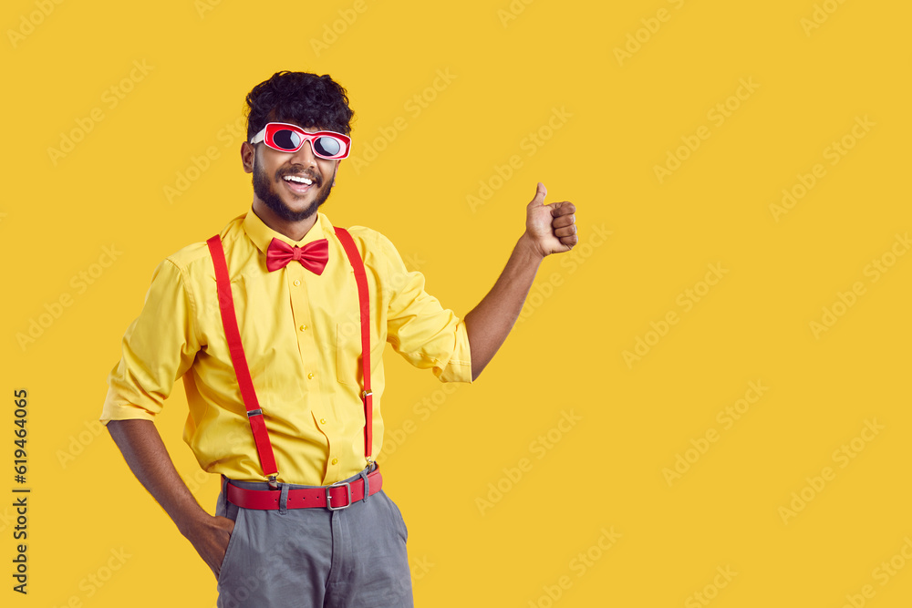 Young contented black haired Indian man in paper funny glasses for party shows thumb up inviting you to entertainment event stands on yellow background with copy space. South Asian portrait Young contented black haired Indian man in paper funny glasses for party shows thumb up inviting you to entertainment event stands on yellow background with copy space. South Asian portrait