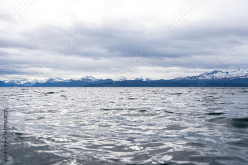 Norwegian fjord with blue water and overcast sky, but in the distance you can see a straight horizon above which dark mountains with white snow open