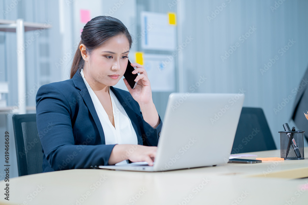 Image of a young beautiful Asian Business woman smiling while working with a laptop and talking to work doing math finance on an office desk, tax, report, accounting, statistics, and research concepts Image of a young beautiful Asian Business woman smiling while working with a laptop and talking to work doing math finance on an office desk, tax, report, accounting, statistics, and research concepts
