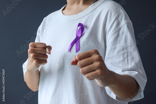 Woman hands in white t-shirt holding purple ribbon gesturing fight cancer against dark grey background. Epilepsy awareness and World Cancer Day concept.  photo