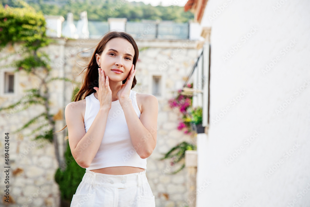 beautiful young brunette with hands on face in white top looking at camera and smiling on white wall background