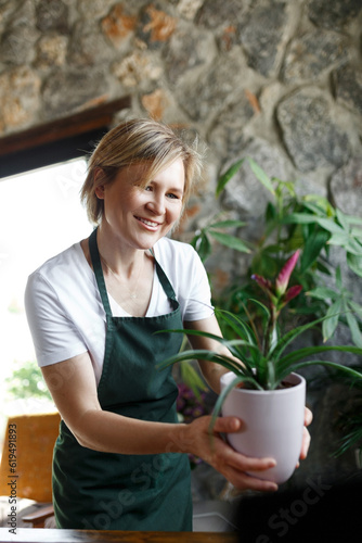 Portrait of smiling young botanist holding a fresh flower plant. Young woman holding small tree in pot in gardening center. Successful botanist and store owner thinking while standing with a pot plant