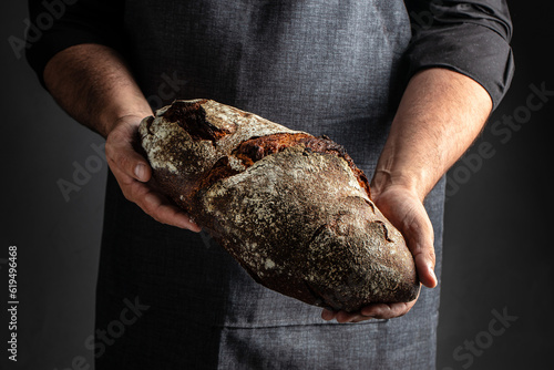 Baker man holding rustic organic loaf of bread in hands on a dark background. banner, menu, recipe place for text, top view
