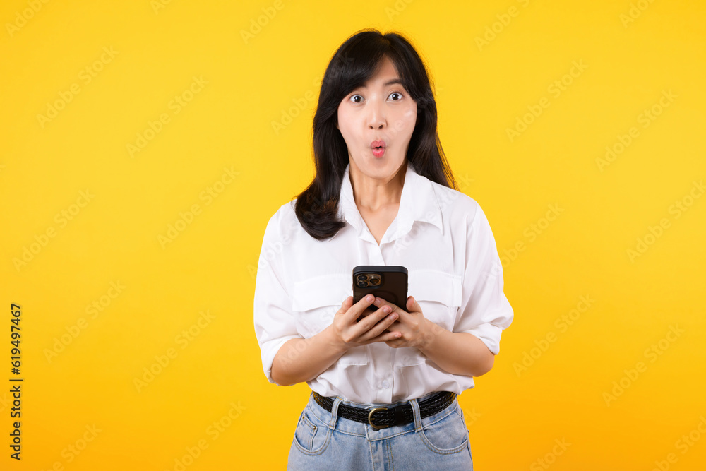 Surprised young asian woman using mobile phone with positive expression, smiles broadly, dressed in white shirt and standing isolated on yellow background. Happy adorable glad woman rejoices success. Surprised young asian woman using mobile phone with positive expression, smiles broadly, dressed in white shirt and standing isolated on yellow background. Happy adorable glad woman rejoices success.
