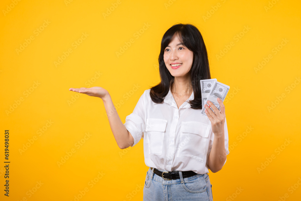 Young happy woman of Asian ethnicity wear white shirt and denim jean holding cash money in dollar and pointing hand to free copy space against yellow background. Investment and financial concept. Young happy woman of Asian ethnicity wear white shirt and denim jean holding cash money in dollar and pointing hand to free copy space against yellow background. Investment and financial concept.