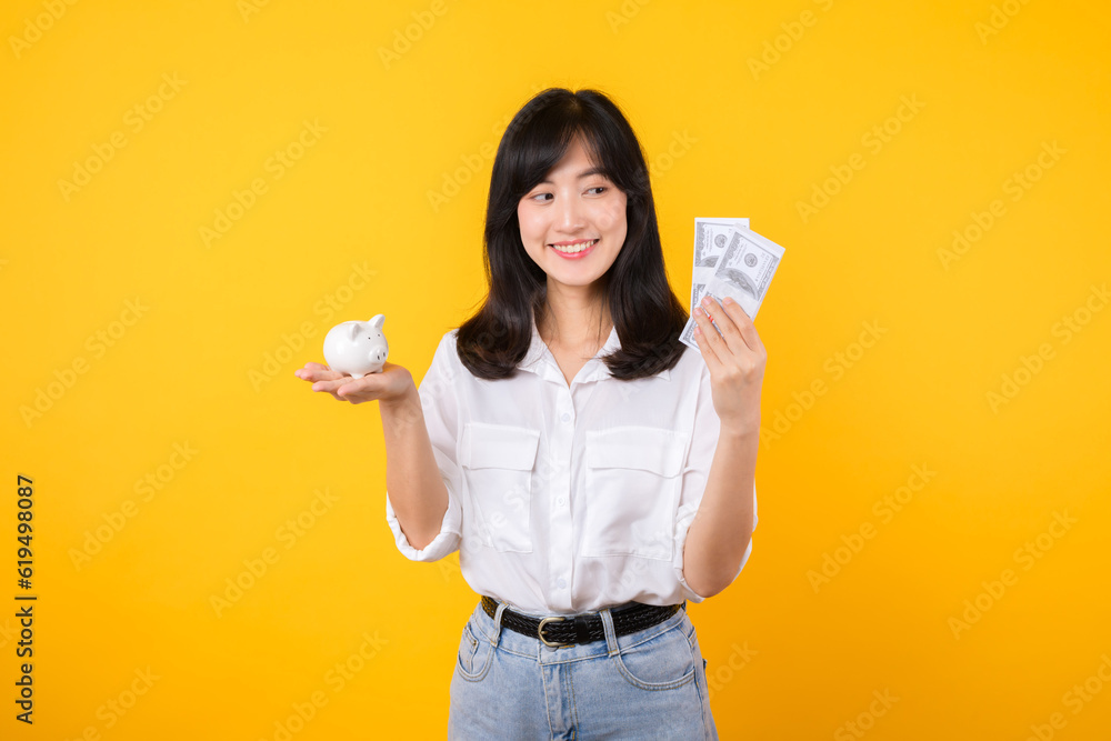Portrait beautiful young business asian woman wearing white shirt and denim jean with a lot of cash money and piggy bank isolated on yellow background. Wealth money saving, finance investment concept. Portrait beautiful young business asian woman wearing white shirt and denim jean with a lot of cash money and piggy bank isolated on yellow background. Wealth money saving, finance investment concept.