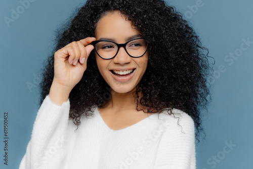 Cheerful woman in glasses, hand on frame, notices awesome thing, wearing white jumper, models on blue background.