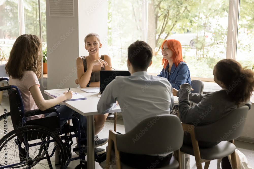 Happy diverse team of students inclusively girl with disability meeting in campus library, sitting at table in library, talking, doing homework, enjoying education, teamwork, communication