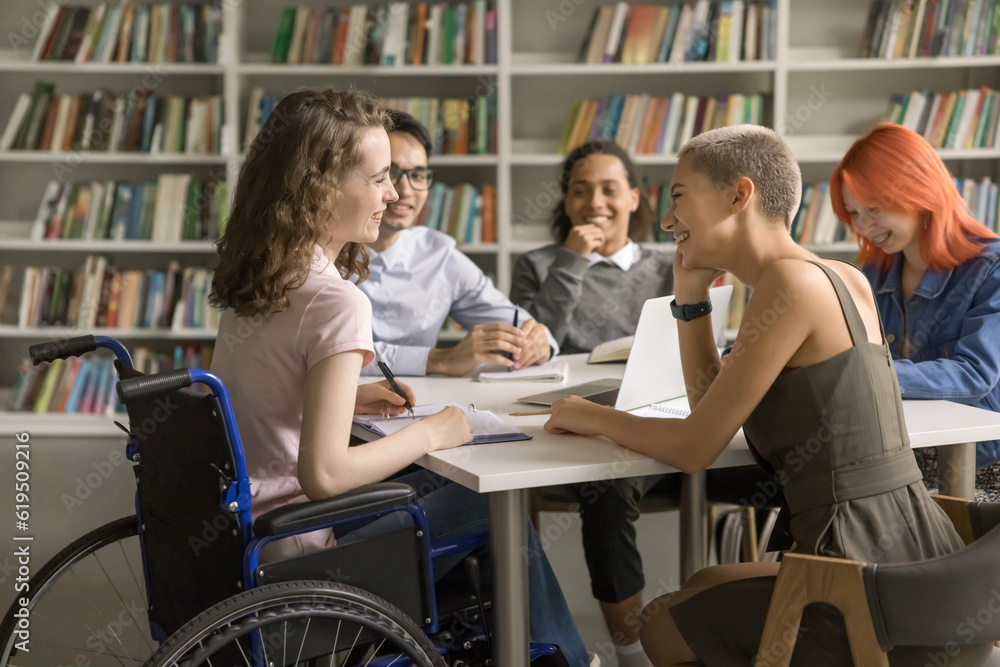 Young student girl with disability cooperating on class project in library with college friends, using wheelchair, sitting at table, talking, chatting with classmates, laughing Young student girl with disability cooperating on class project in library with college friends, using wheelchair, sitting at table, talking, chatting with classmates, laughing