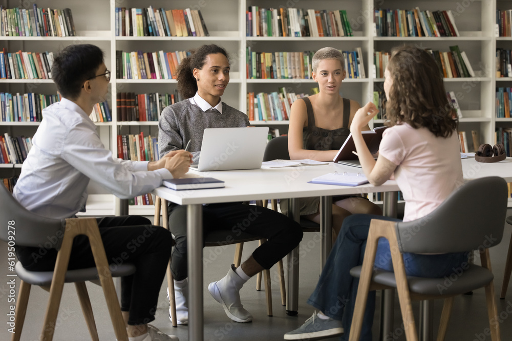 Diverse group of fresh students studying together in university campus, sitting at table in library, talking, discussing education, class project, working on home assignment in multiethnic team