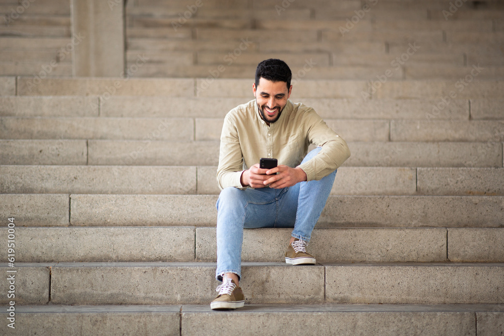 Young arab man sitting on steps looking at his mobile phone and smiling Young arab man sitting on steps looking at his mobile phone and smiling