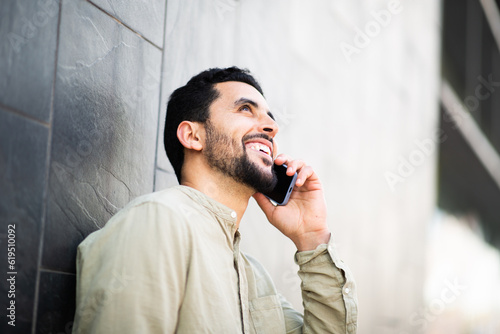 Smiling young arab guy talking on cell phone leaning to a wall