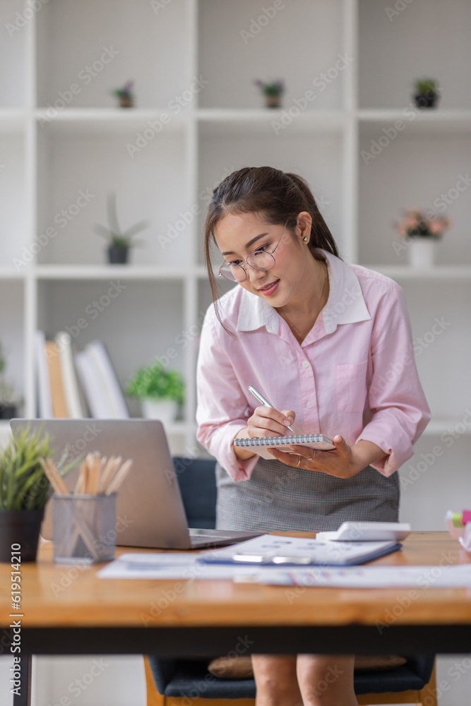 Asian businesswoman working on a laptop computer at her desk in a bright modern office, doing calculating expense financial report finance making notes paper graph data document. Asian businesswoman working on a laptop computer at her desk in a bright modern office, doing calculating expense financial report finance making notes paper graph data document.