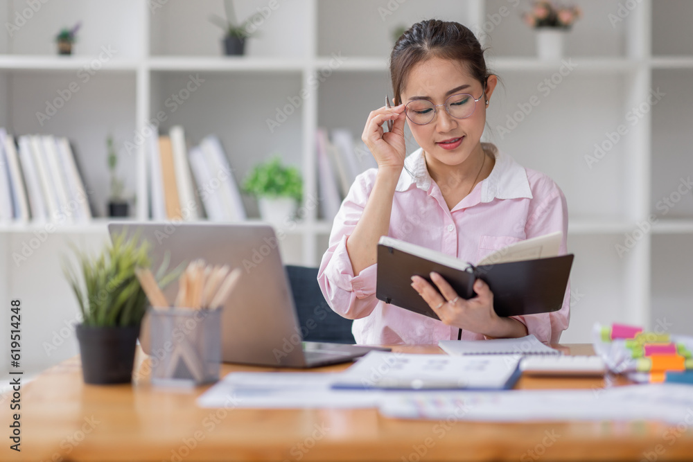 Asian businesswoman working on a laptop computer at her desk in a bright modern office, doing calculating expense financial report finance making notes paper graph data document. Asian businesswoman working on a laptop computer at her desk in a bright modern office, doing calculating expense financial report finance making notes paper graph data document.