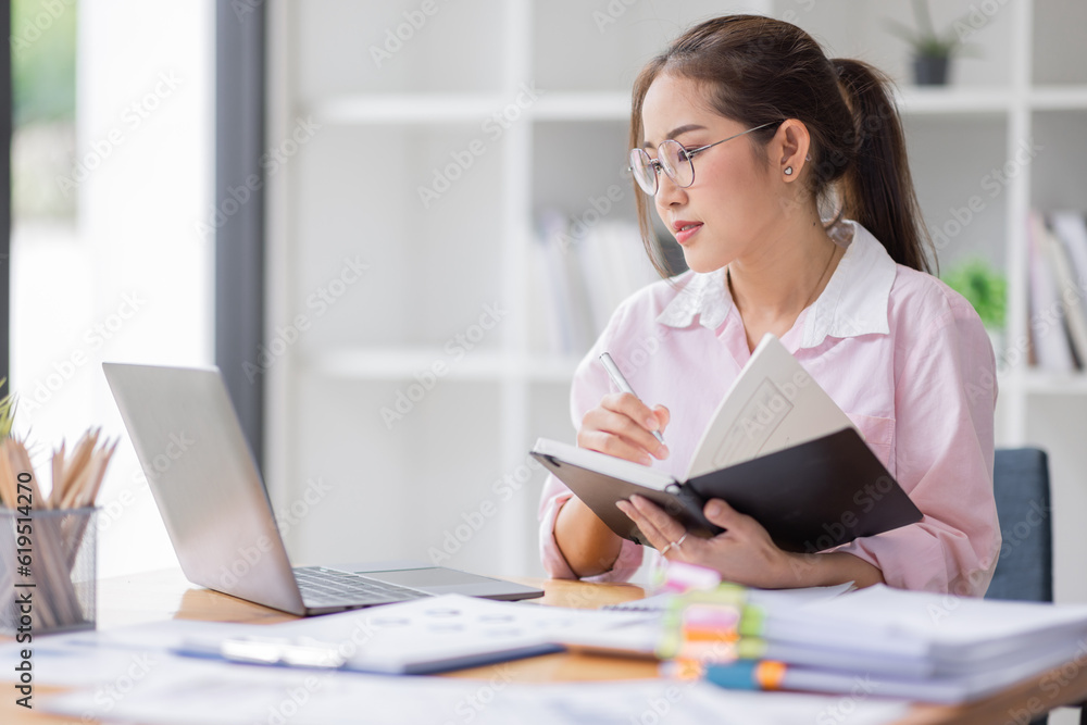 Asian businesswoman working on a laptop computer at her desk in a bright modern office, doing calculating expense financial report finance making notes paper graph data document. Asian businesswoman working on a laptop computer at her desk in a bright modern office, doing calculating expense financial report finance making notes paper graph data document.