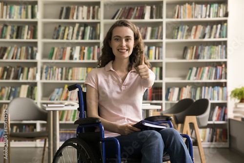 Happy pretty student girl using wheelchair, posing in library, showing like thumb up hand with toothy smile, celebrating educational success, achievement, enjoying inclusive environment