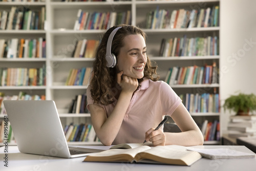 Happy college student girl in wireless headphones enjoying studying in library, sitting at laptop, open books, working on essay, article, writing notes, looking away, thinking, smiling, laughing