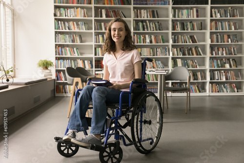 Cheerful young college student with disability using wheelchair, visiting campus library, holding book, looking at camera with toothy smile, promoting inclusive educational environment
