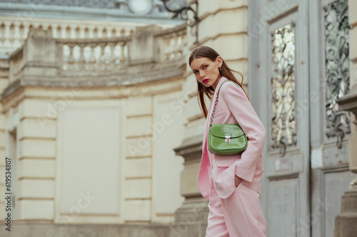 Fashionable elegant confident woman wearing trendy pink suit blazer, wide trousers, with green faux leather shoulder bag, walking in street. Outdoor fashion portrait. Copy, empty space for text