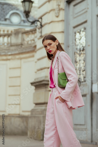 Fashionable elegant confident woman wearing trendy pink suit blazer, wide trousers, with green faux leather shoulder bag, walking in street. Outdoor fashion portrait