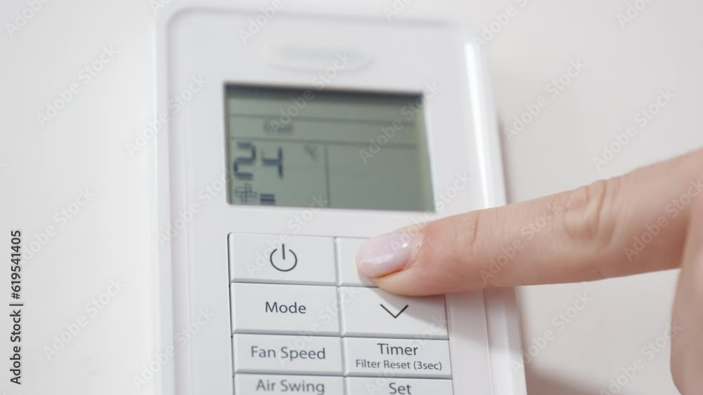 The camera focuses on the air conditioner remote control on the white wall as the woman turns it on and increases the temperature to the maximum. The camera focuses on the air conditioner remote control on the white wall as the woman turns it on and increases the temperature to the maximum.