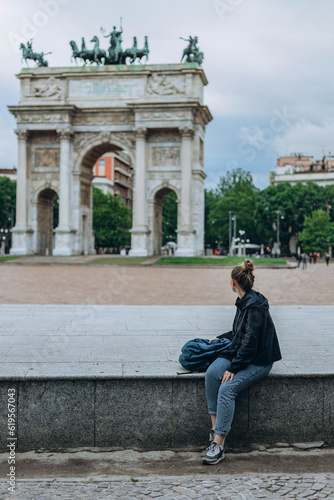 Milan, Italy- May 13, 2023: Girl sits near Arch of Peace or Sempione Gate in Milan, near Sempione Park. Exterior view of the marble gate of the Arco della Sempione, a peace memorial in Milan.