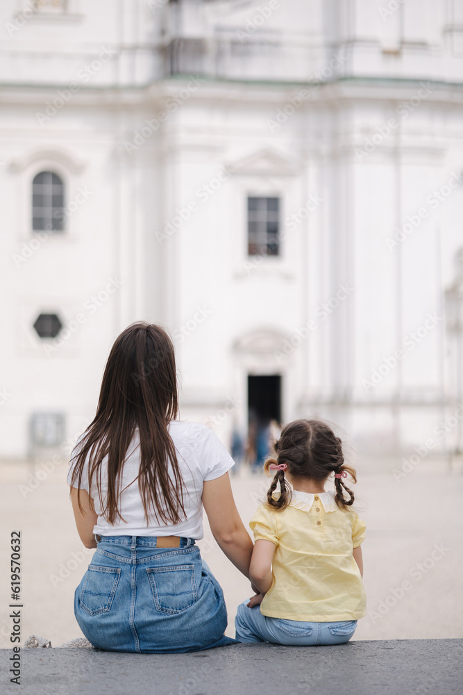 Back view of little girl sits with her mom on stone bench outdoor Back view of little girl sits with her mom on stone bench outdoor