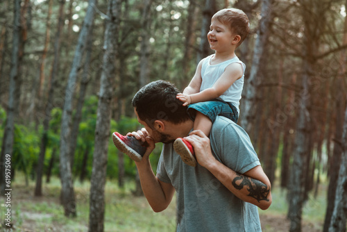 father's day. Happy European-looking father walks with his young cute two-year-old son on his shoulders in the woods