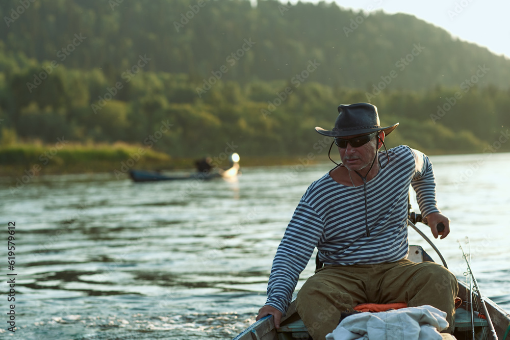 A bruA brutal man, a fisherman aged 60 plus in a vest and a black hat, drives a motor boat on the river. Sunset time on the river. The concept of tourism and recreation. High resolution photo