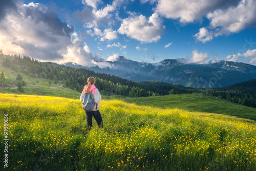 Fototapeta Girl on the hill with yellow flowers and green grass in beautiful alpine mountain valley at sunset in summer