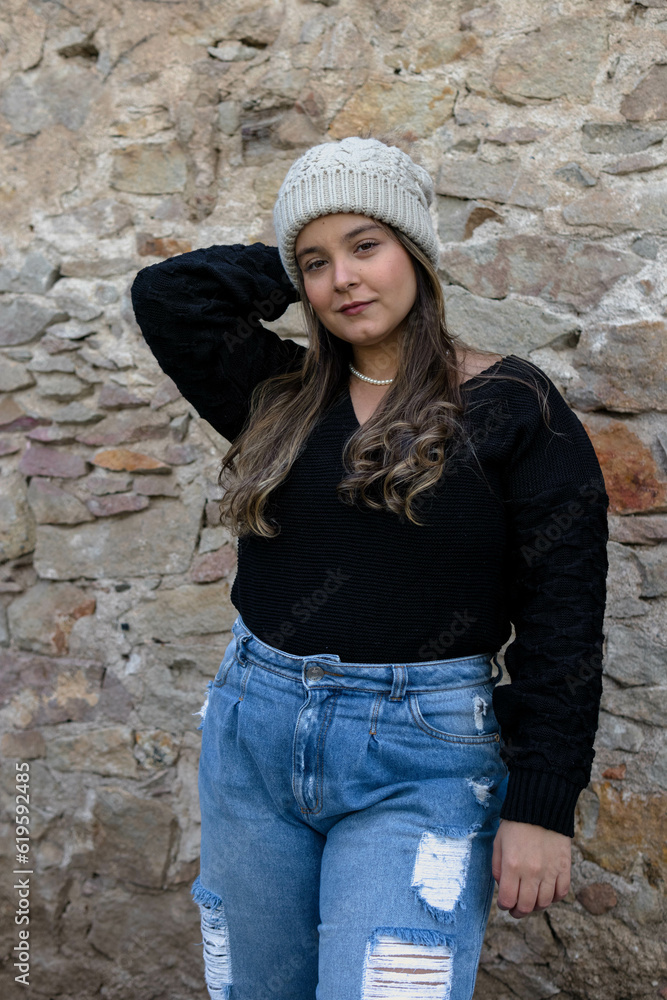 three quarter lenght image of apretty young woman with latin features standing ahead of a stone wall with one hand on her hair