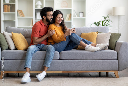 Happy indian man and woman using smartphone while relaxing on couch together