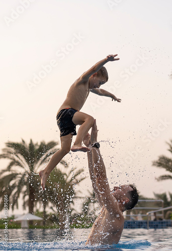Dad and son are playing in the swimming pool. The father tosses the son up.