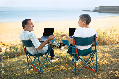 European Senior Spouses Using Laptop Sitting At Sea Beach Outdoor